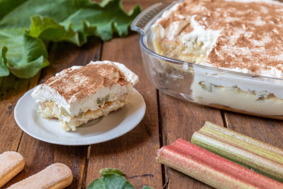 Eine Portion Rhabarber-Tiramisu auf einem Teller. Das restliche Dessert steht in einer Auflaufform dahinter.