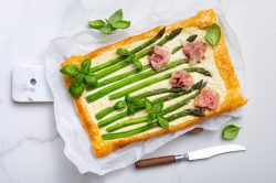 Spargel-Blätterteig-Pizza mit Schinken und Basilikum, daneben ein Messer, Draufsicht.