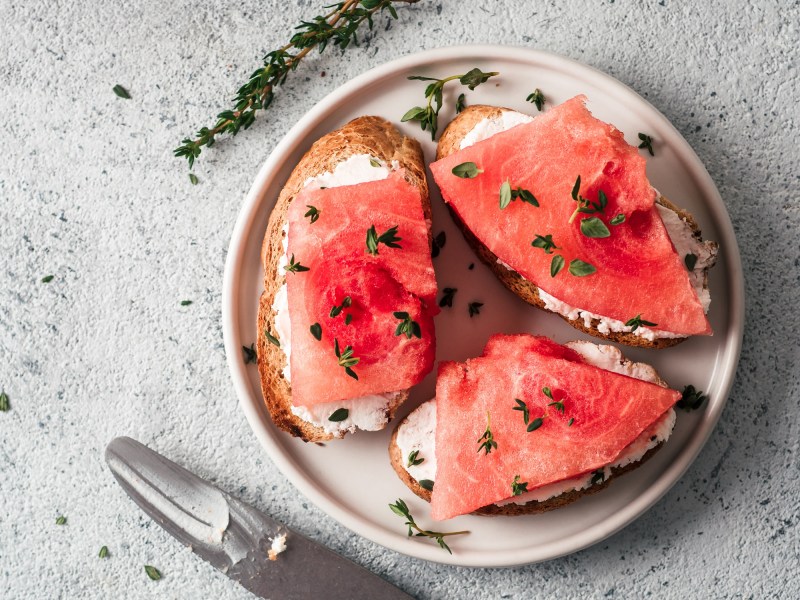 Drei Brote mit veganem Thunfisch aus Wassermelone auf einem Teller, garniert mit Kräutern, daneben ein Messer, Draufsicht.