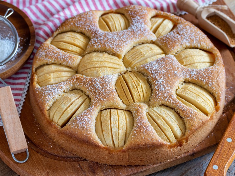 Versunkener Apfelkuchen mit Zimtäpfeln bestreut mit Puderzucker, daneben ein Sieb. Nahaufnahme.