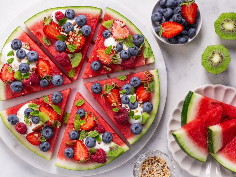 Eine Wassermelonen-Pizza auf einem Teller Melonenstücke, Kiwi und Beeren daneben auf einem Tisch