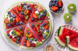 Eine Wassermelonen-Pizza auf einem Teller Melonenstücke, Kiwi und Beeren daneben auf einem Tisch