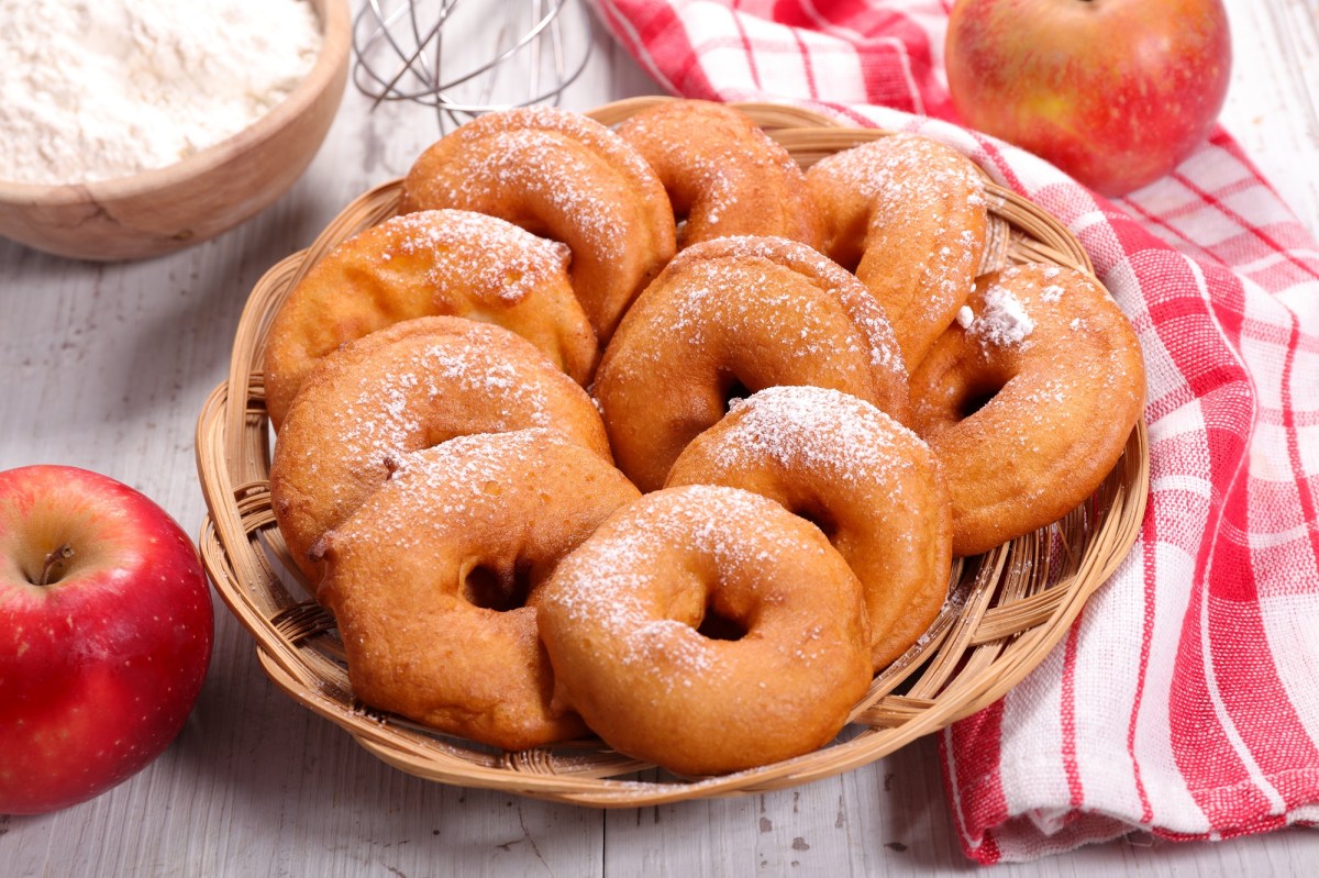 Apfel-Beignets, mit Puderzucker bestreut, in einem Korb. Daneben liegen Äpfel.