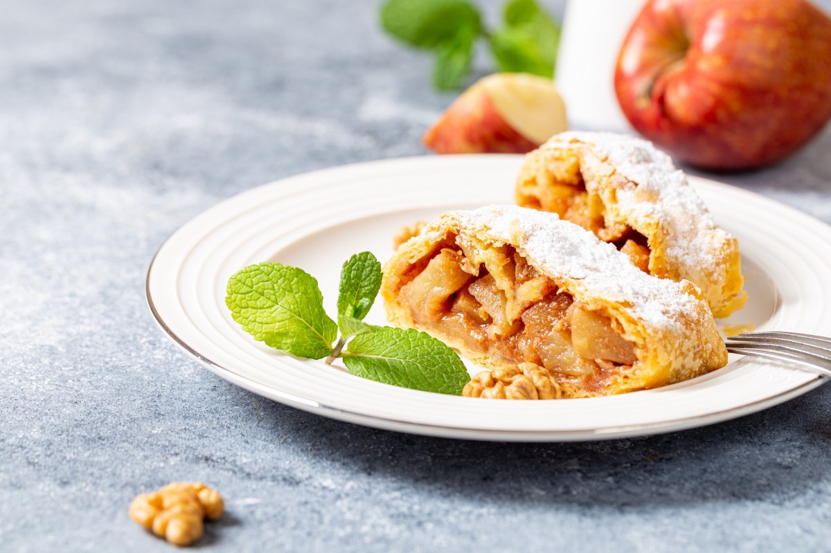 Zwei Stücke Apfelstrudel mit Blätterteig auf einem Teller, bestäubt mit Puderzucker, im Hintergrund Äpfel.