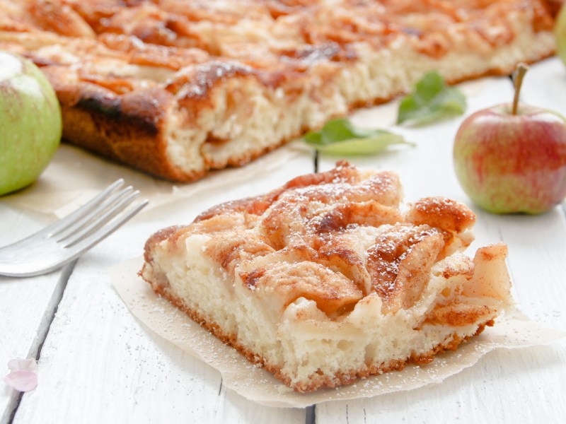 Blechkuchen-Rezepte: ein Stück Apfelkuchen vom Blech auf einem Stück Backpapier, im Hintergrund der Kuchen und 2 Äpfel.