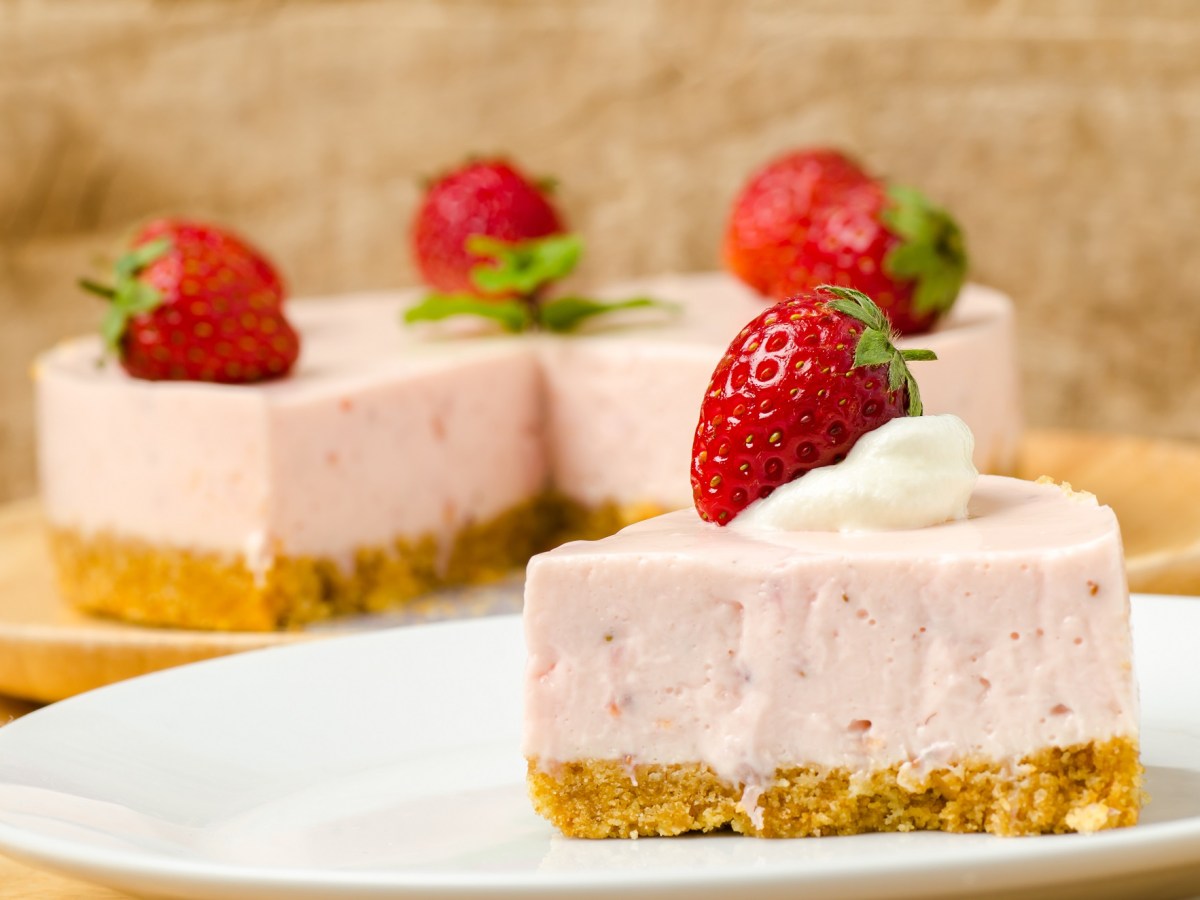 Ein Stück Erdbeermousse-Kuchen, garniert mit einer Erdbeere, auf einem Teller, im Hintergrund der angeschnittene Kuchen.