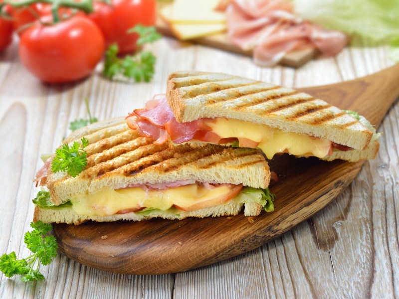 Zwei gegrillte Käse-Schinken-Sandwiches auf einem Brett, im Hintergrund frische Tomaten.