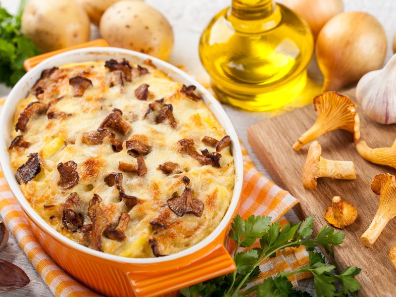 Kartoffelauflauf mit Pfifferlingen mit einer Auflaufform, daneben Zutaten wie Pfifferlinge, Öl und Zwiebeln.
