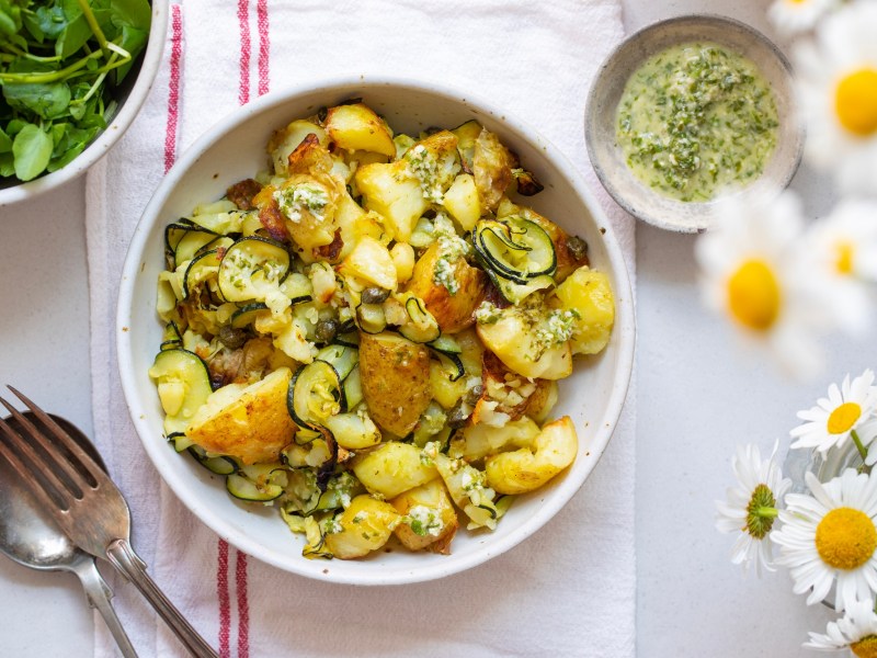 Kartoffelsalat mit gegrillter Zucchini in einer weißen Schale. Daneben steht eine Schale mit Dressing.