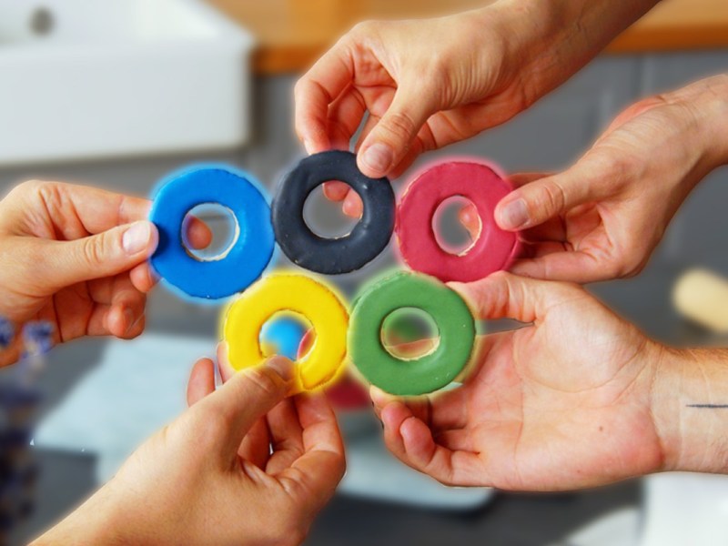 Fünf Hände halten bunte Olympiade-Kekse zu den olympischen Ringen.