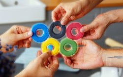 Fünf Hände halten bunte Olympiade-Kekse zu den olympischen Ringen.