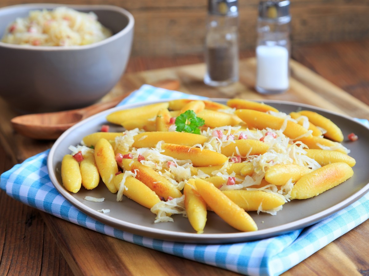 Ein Teller Schupfnudeln mit Sauerkraut und Speck, im Hintergrund eine Schüssel Sauerkraut sowie Salz- und Pfefferstreuer.