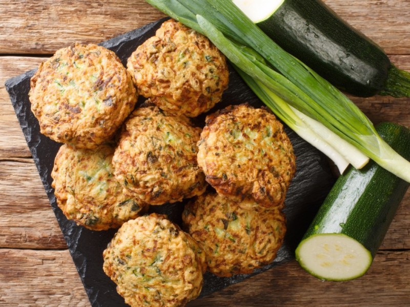 Mehrere Zucchini-Bällchen in der Draufsicht. Daneben liegen Zucchini und Frühlingszwiebeln.