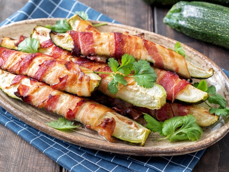 Zucchini im Speckmantel auf einem ovalen Teller, daneben eine Zucchini.