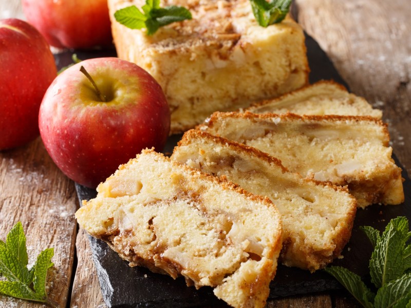 angeschnittener Apfelkuchen aus der Kastenform, daneben ganze Äpfel.