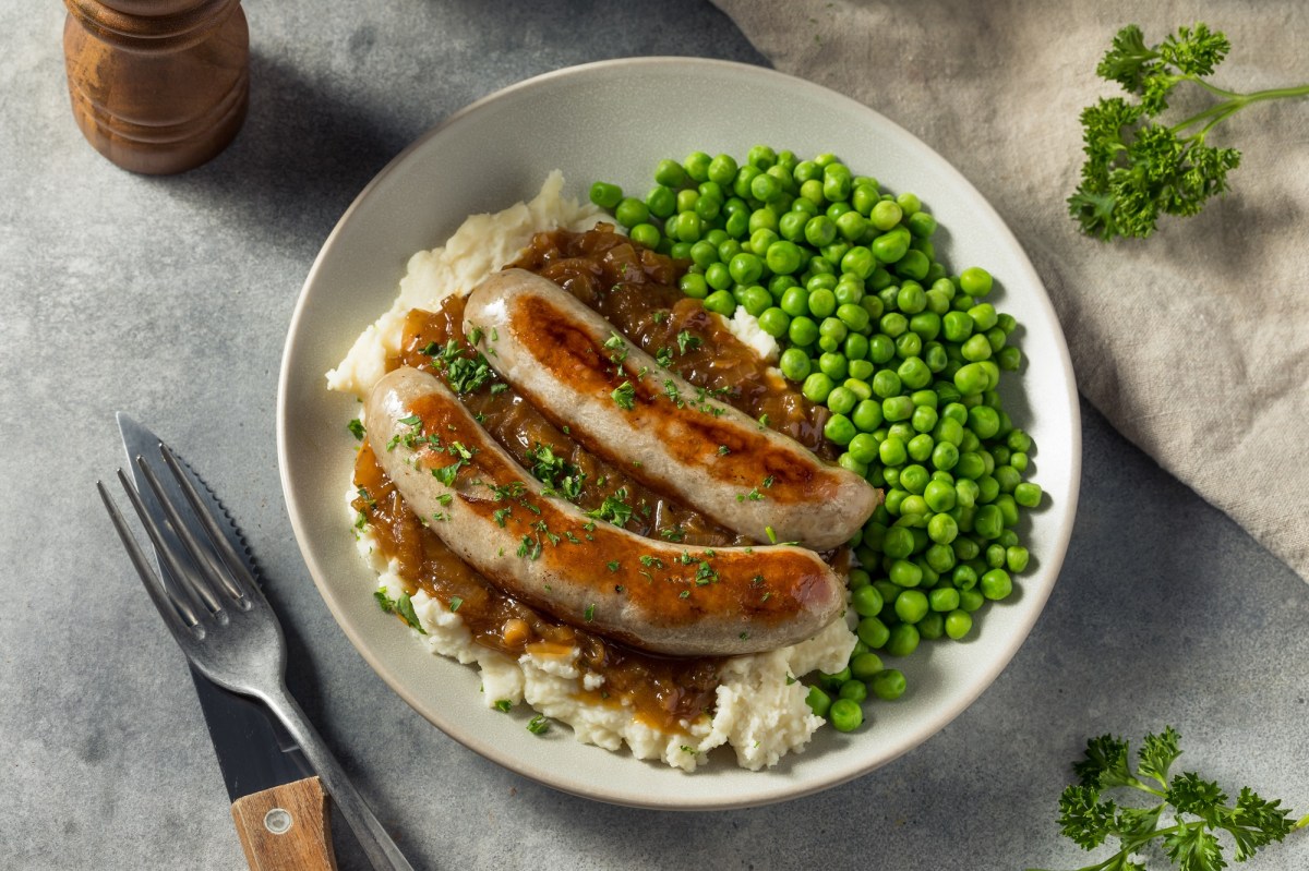 Bangers and Mash: ein Teller mit zwei Bratwürsten, Kartoffelbrei, Erbsen und brauner Soße, daneben Messer und Gabel, Draufsicht.