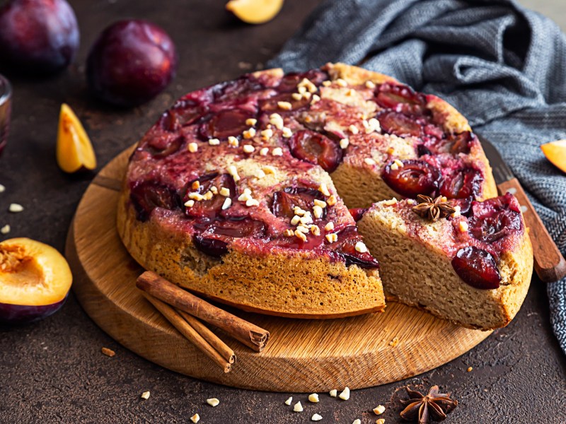 Gestürzter Pflaumenkuchen auf einem Holzbrett. Ein Stück wurde bereits abgeschnitten.