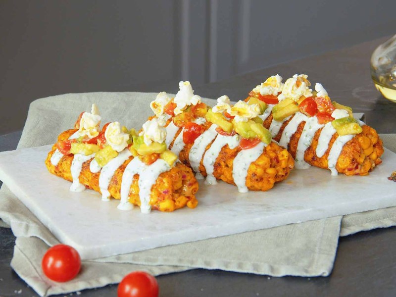 3 Käse-Mais-Kroketten auf einem Teller, garniert mit Sour Cream, Tomaten-Avocado-Salat und Popcorm.