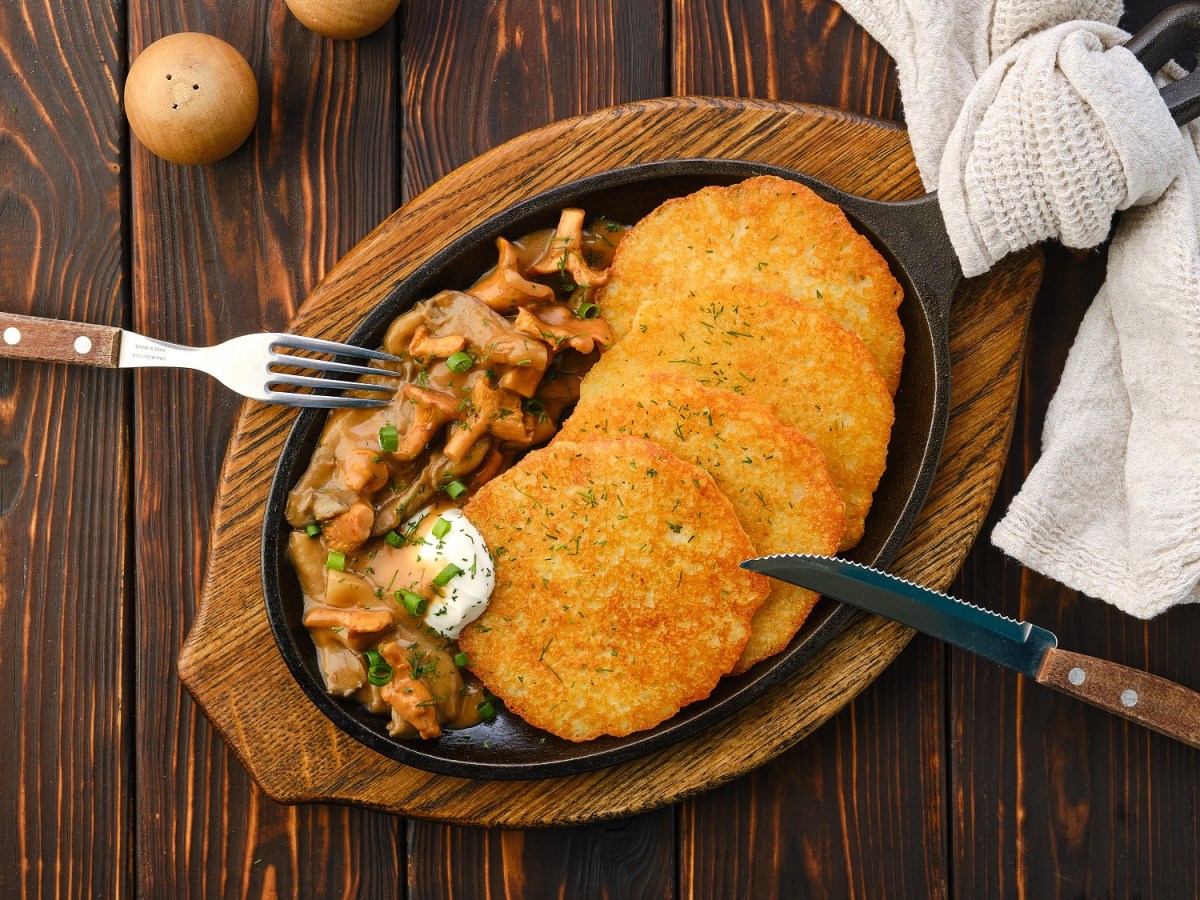 Eine ovale gusseiserne Pfanne mit Kartoffelpuffer mit Pfifferling-Rahmsoße, daneben Messer und Gabel. Draufsicht.