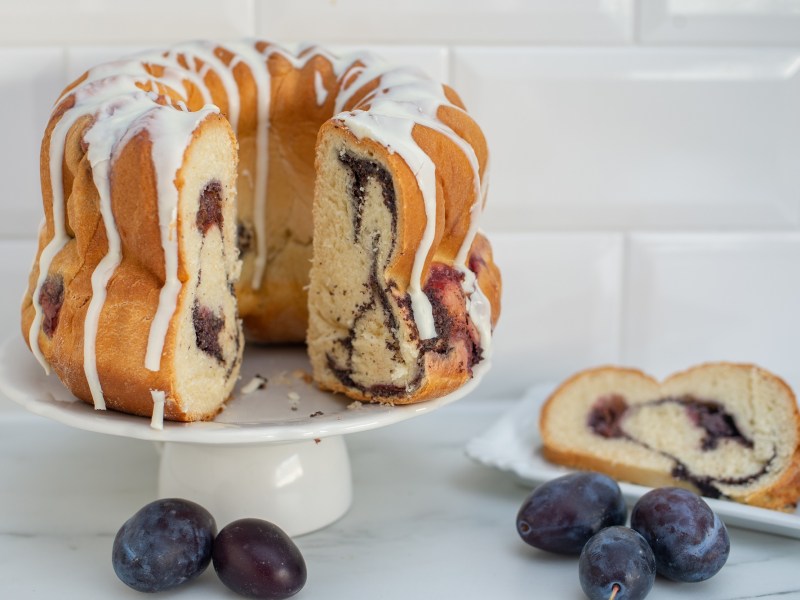 Ein angeschnittener Marmorkuchen mit Pflaumen in Gugelhupf-Form, daneben ein Teller mit einem Stück sowie frische Pflaumen.