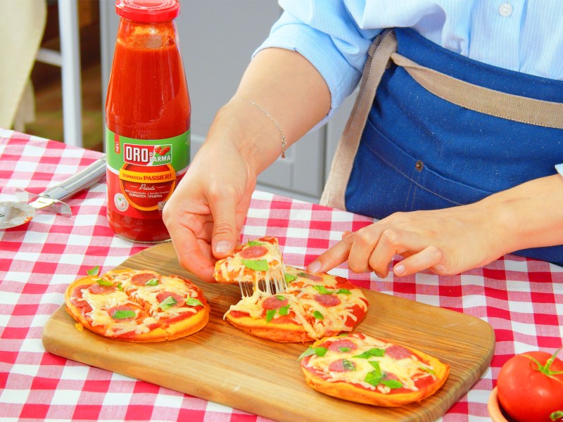 Mini-Pizzen mit Tomatenteig, Käse und Salami auf einem Brett, ein Stück wird herausgenommen.