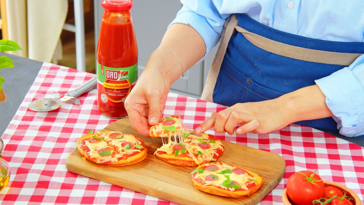 Mini-Pizzen mit Tomatenteig, Käse und Salami auf einem Brett, ein Stück wird herausgenommen.