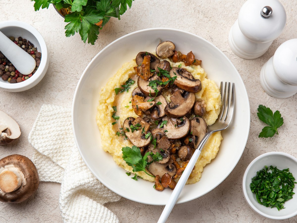 Polenta mit Pilzen auf einem weißen Teller. Daneben liegen die Zutaten und Besteck.