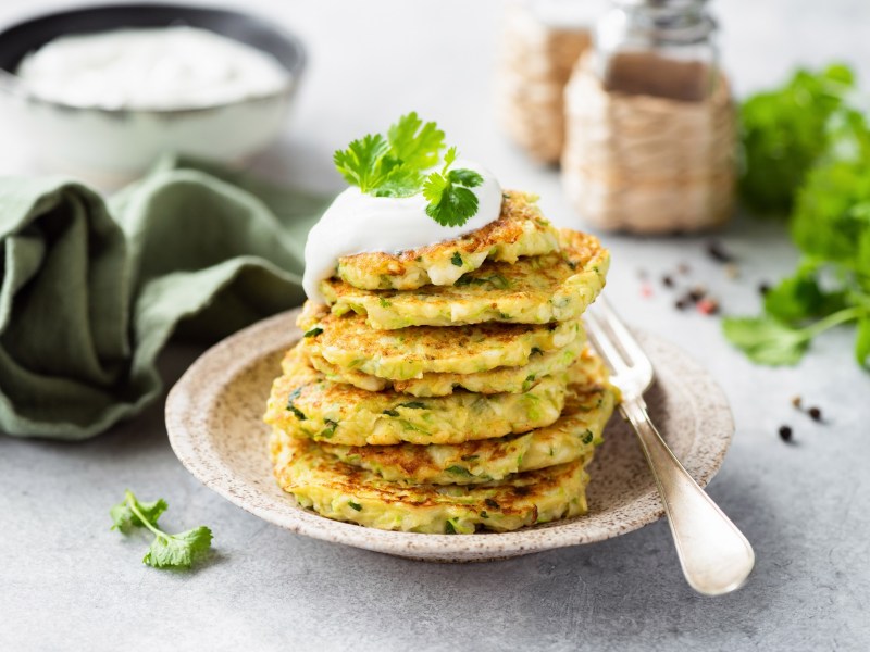 Ein Stapel Zucchini-Pancakes mit Hüttenkäse auf einem weißen Teller. Daneben liegt eine Gabel. Die Pancakes sind mit Joghurt und Petersilie garniert.