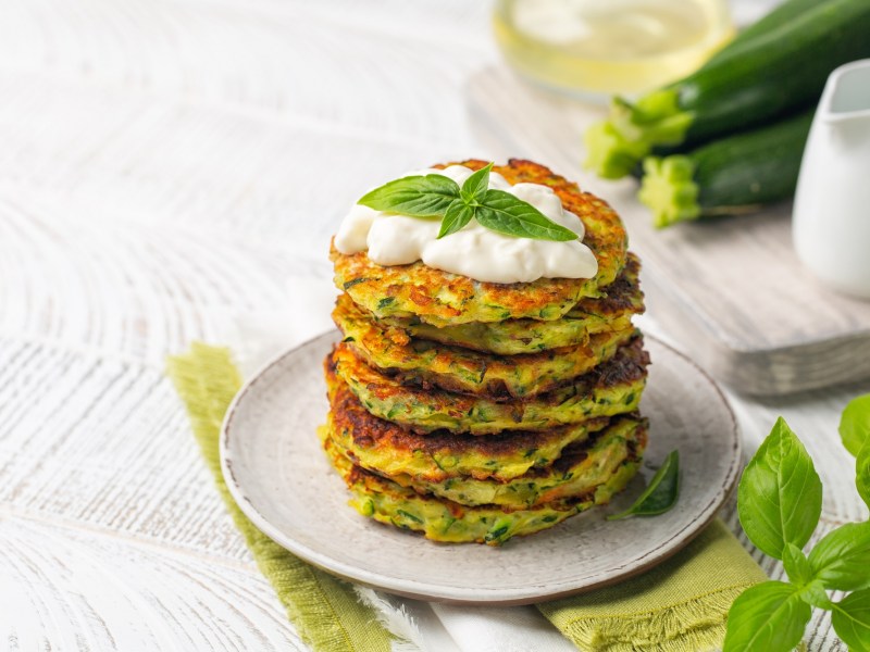 Ein Stapel Zucchini-Parmesan-Puffer auf einem Teller, garniert mit einem Klecks Schmand und Basilikum.