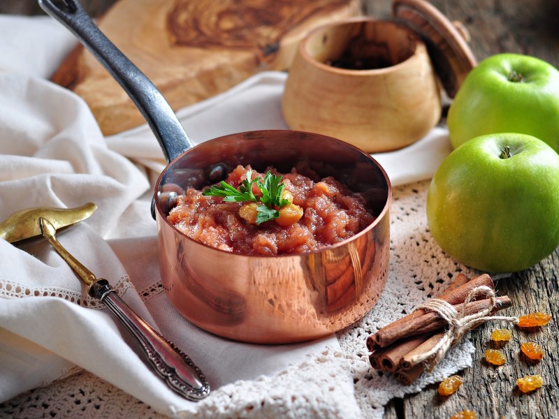 Ein kleiner Topf mit Apfelchutney und Rosinen, drumherum frische grüne Äpfel, Sultaninen und Zubehör.