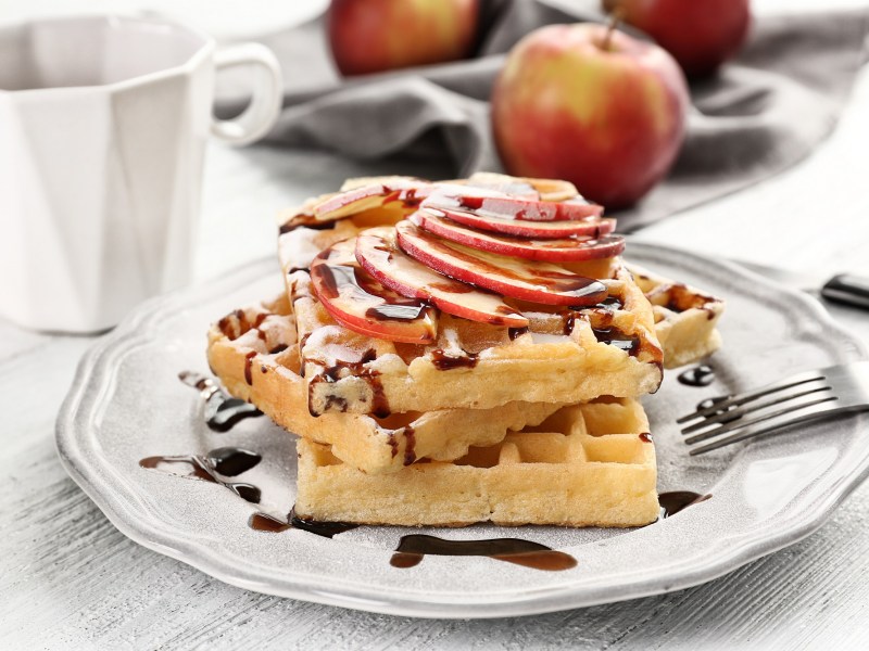 Apfelwaffeln mit Schokosoße und Apfelscheiben auf einem weißen Teller. Dahinter liegt ein Apfel.