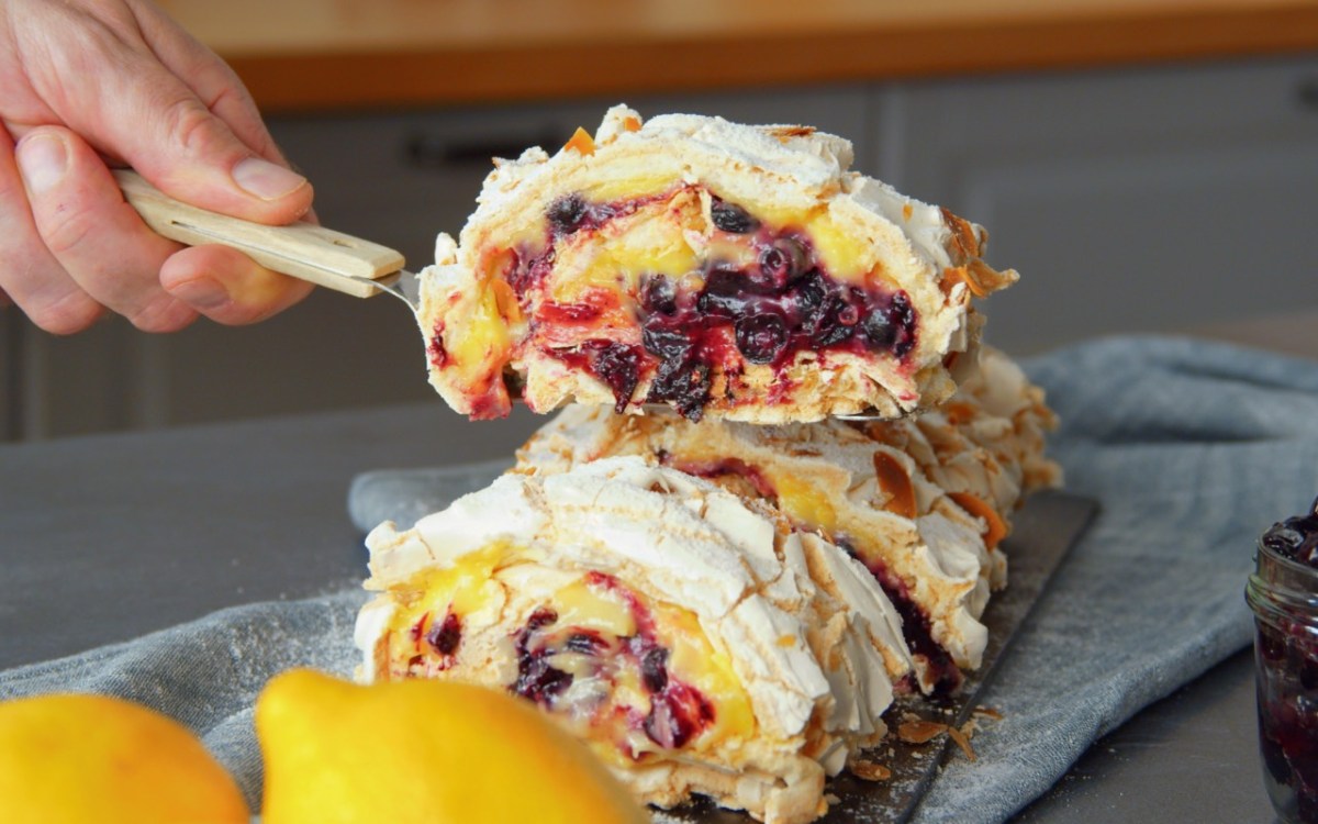 Ein Stück gefüllte Baiserrolle mit Lemon Curd und Pflaumenkompott wird mit einem Tortenheber entnommen.