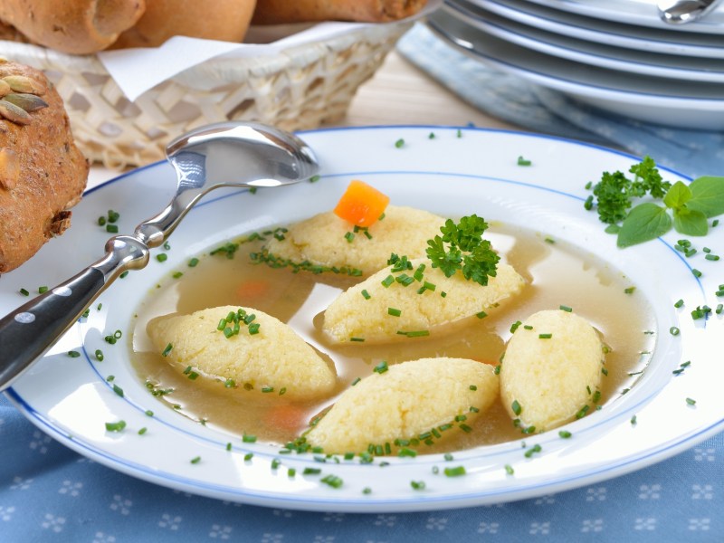 Grießschöberl-Suppe auf einem Teller, daneben ein Brötchen. Nahaufnahme.