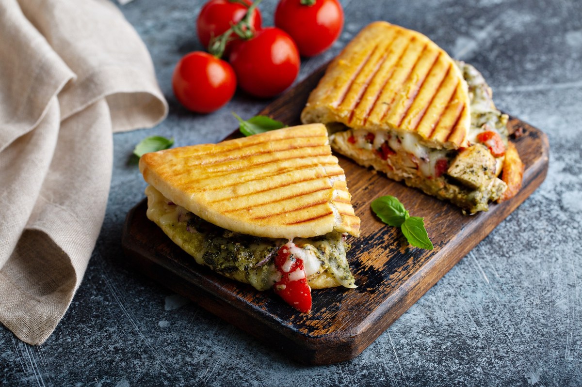 2 Italienische Hähnchen-Paninis mit grünem Pesto und Mozzarella auf einem Brett, im Hintergrund Tomaten.