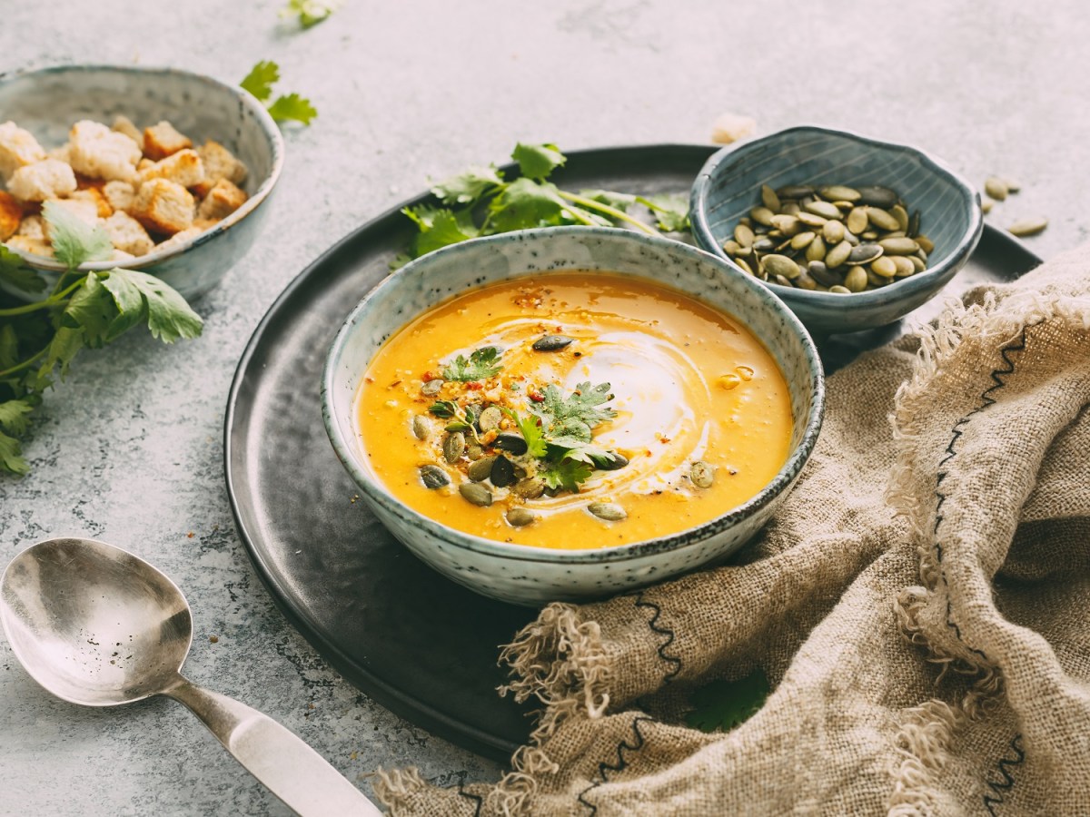 Eine Schüssel Kürbis-Curry-Suppe mit Kernen, Croûtons und Kräutern verfeinert, daneben je eine Schüssel mit Kernen und Croûtons.