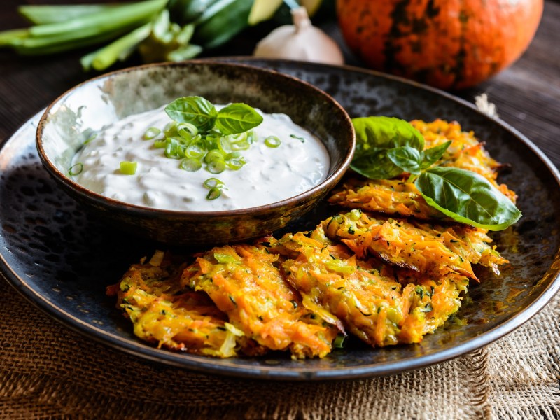 Ein Teller Kürbis-Kartoffel-Rösti und eine Schale weißer Dip, dahinter ein Kürbis und Frühlingszwiebeln.