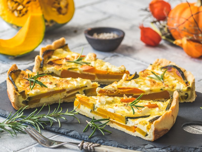 4 Stücke Kürbis-Quiche mit Tofu auf einer Schieferplatte, im Hintergrund verschiedene Kürbisse.