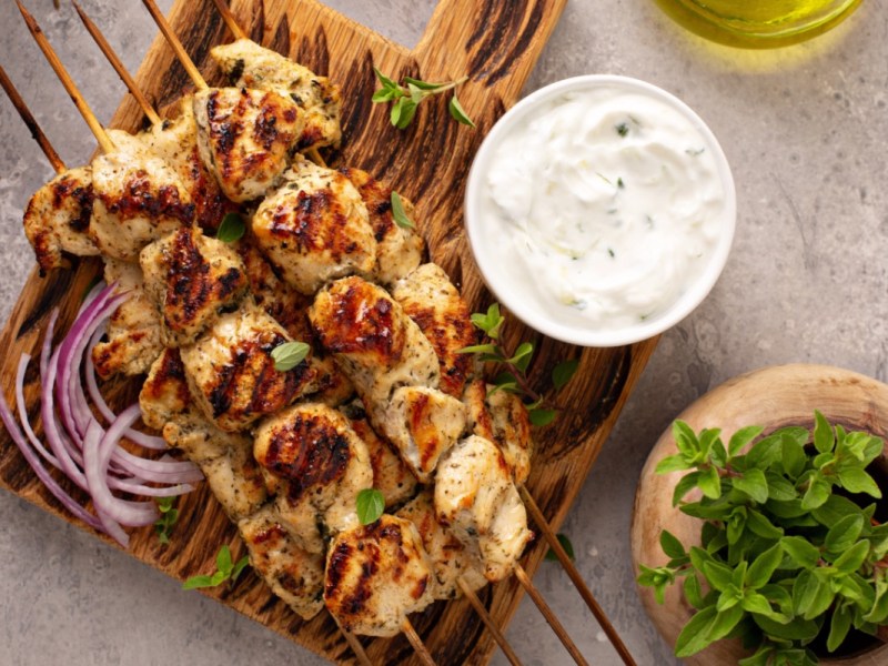 Mehrere Souvlaki. Daneben Zwiebeln und eine Schale mit Zaziki.