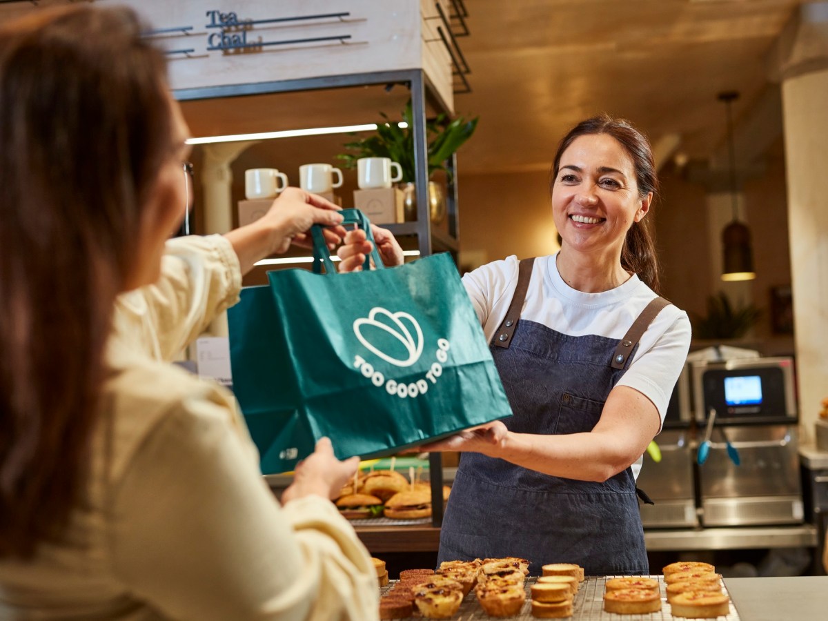 Eine dunkelhaarige Frau reicht einer anderen in einem Café eine Tüte mit der Aufschrit "Too Good To Go" über den Tresen.