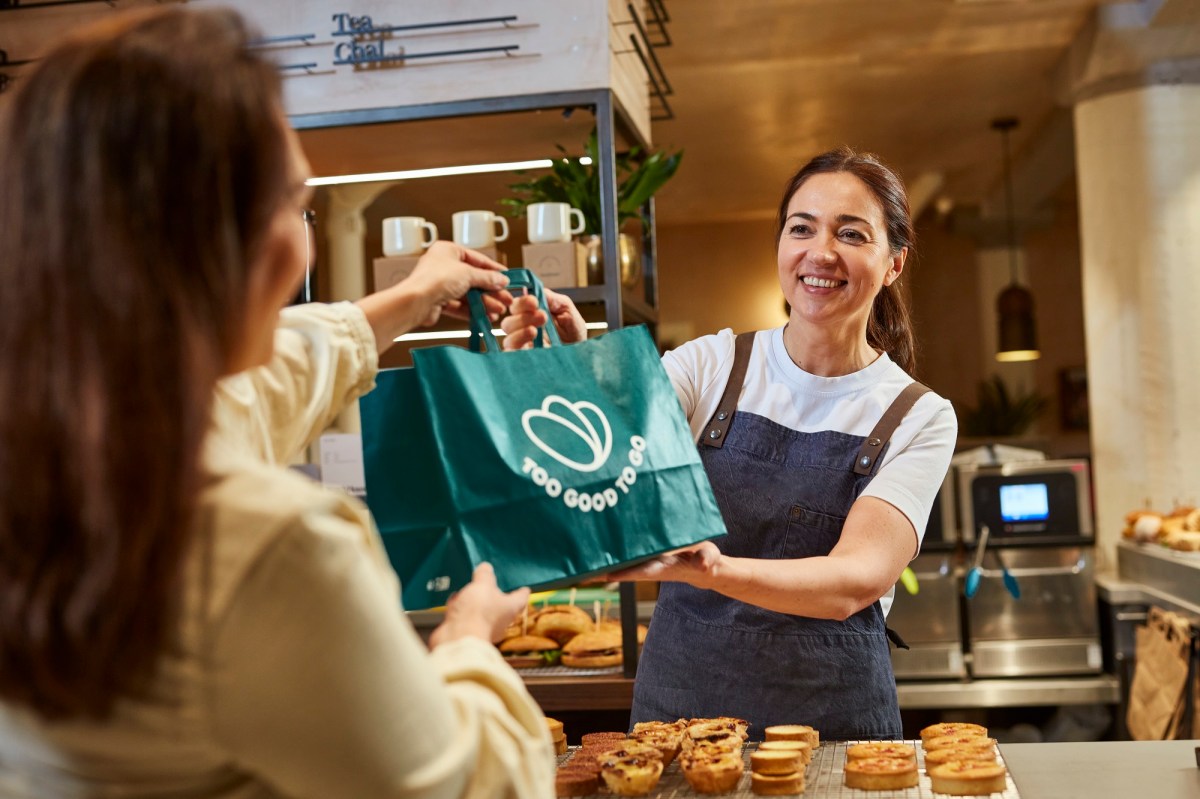 Eine dunkelhaarige Frau reicht einer anderen in einem Café eine Tüte mit der Aufschrit "Too Good To Go" über den Tresen.