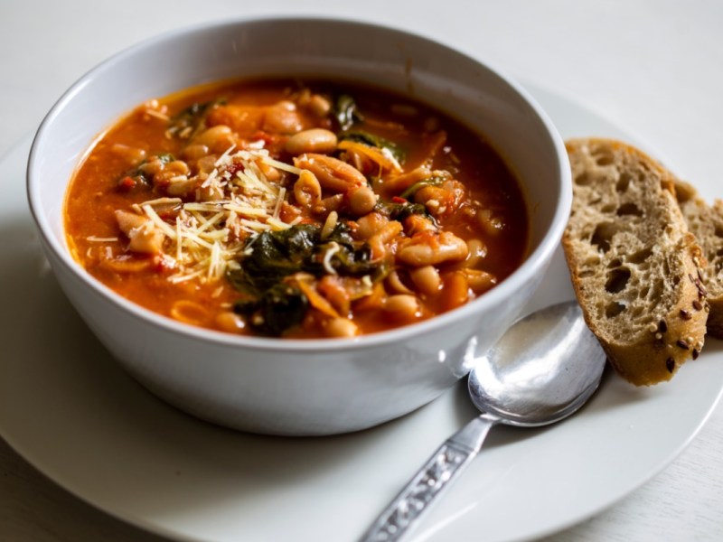 Eine Schüssel mit toskanischer Bohnensuppe. Daneben liegen eine Scheibe Brot und Löffel.