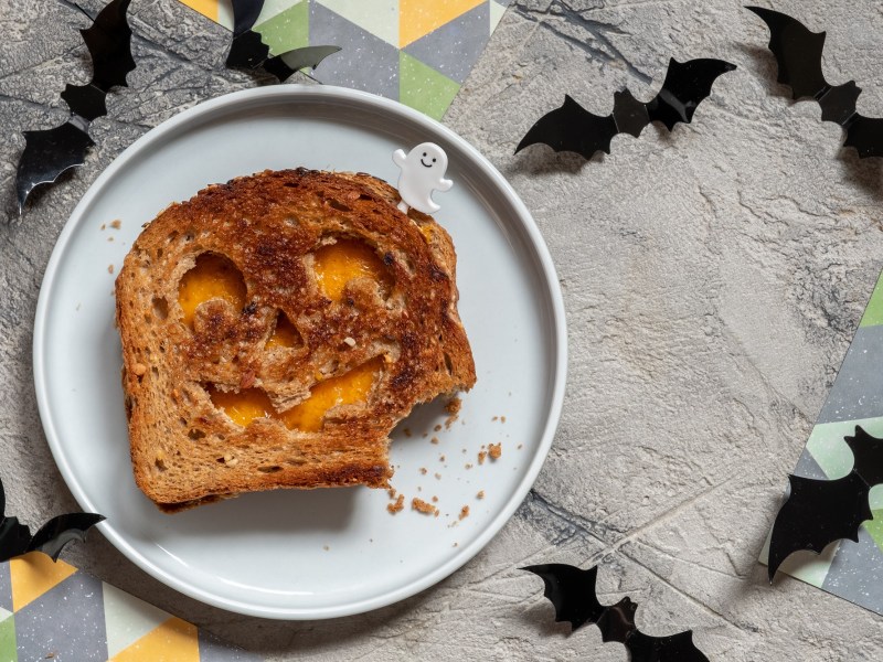 Halloween-Sandwich mit Käse und Kürbisgesicht auf einem Teller in der Draufsicht, drumherum Halloween-Deko.