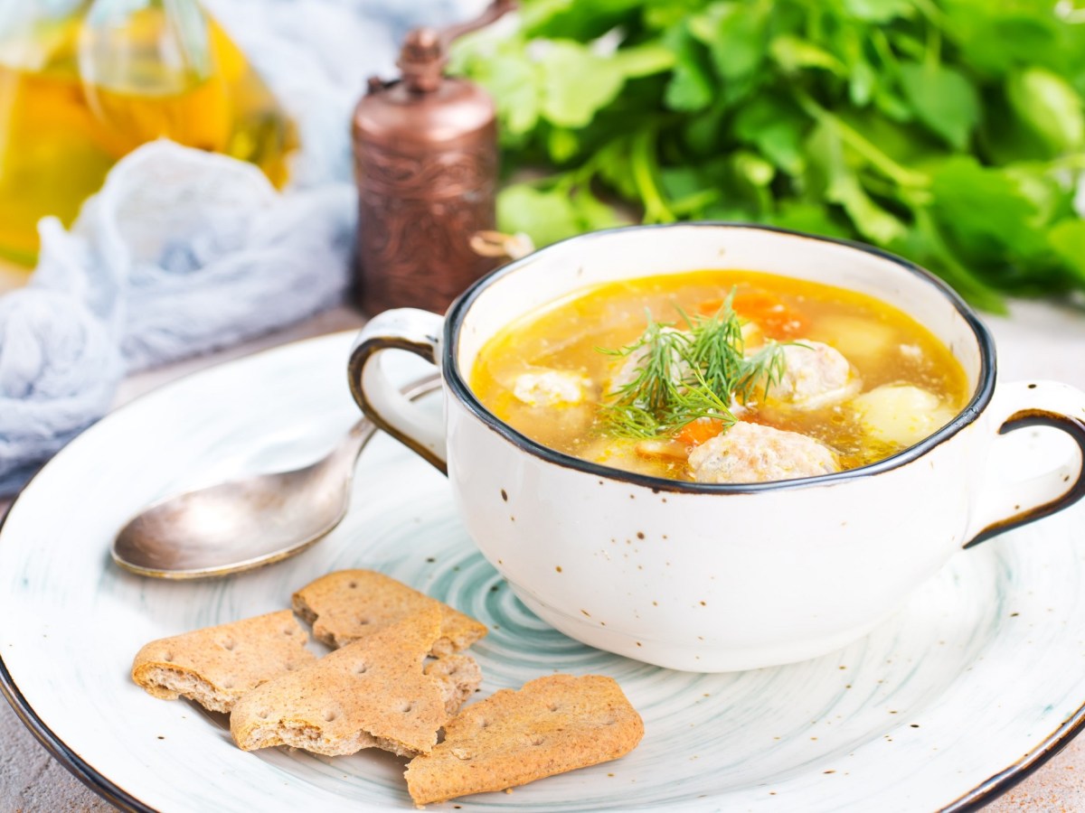 Eine Schüssel Hochzeitssuppe mit Klößen und Gemüse auf einem Teller, daneben ein Löffel und Cracker, im Hintergrund Kräuter.