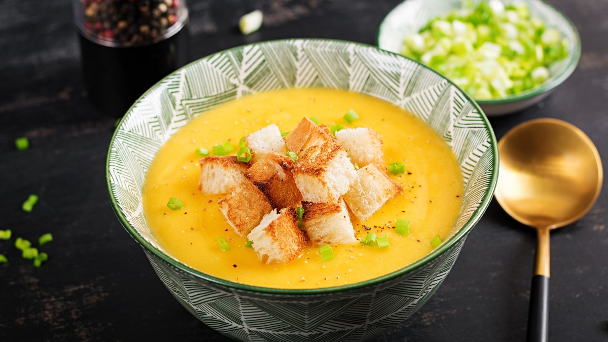 Eine Schüssel Kürbis-Kartoffel-Suppe mit Croûtons und Frühlingszwiebel, daneben ein Löffel und eine Schale Frühlingszwiebel.