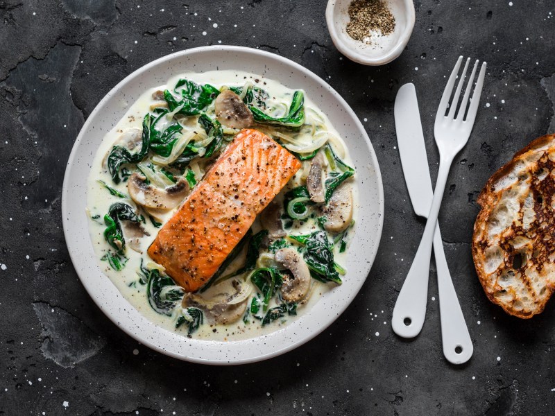 Lachs mit Spinat und Pilzen auf einem weißen Teller. Daneben liegt Besteck und eine Scheibe geröstetes Brot.