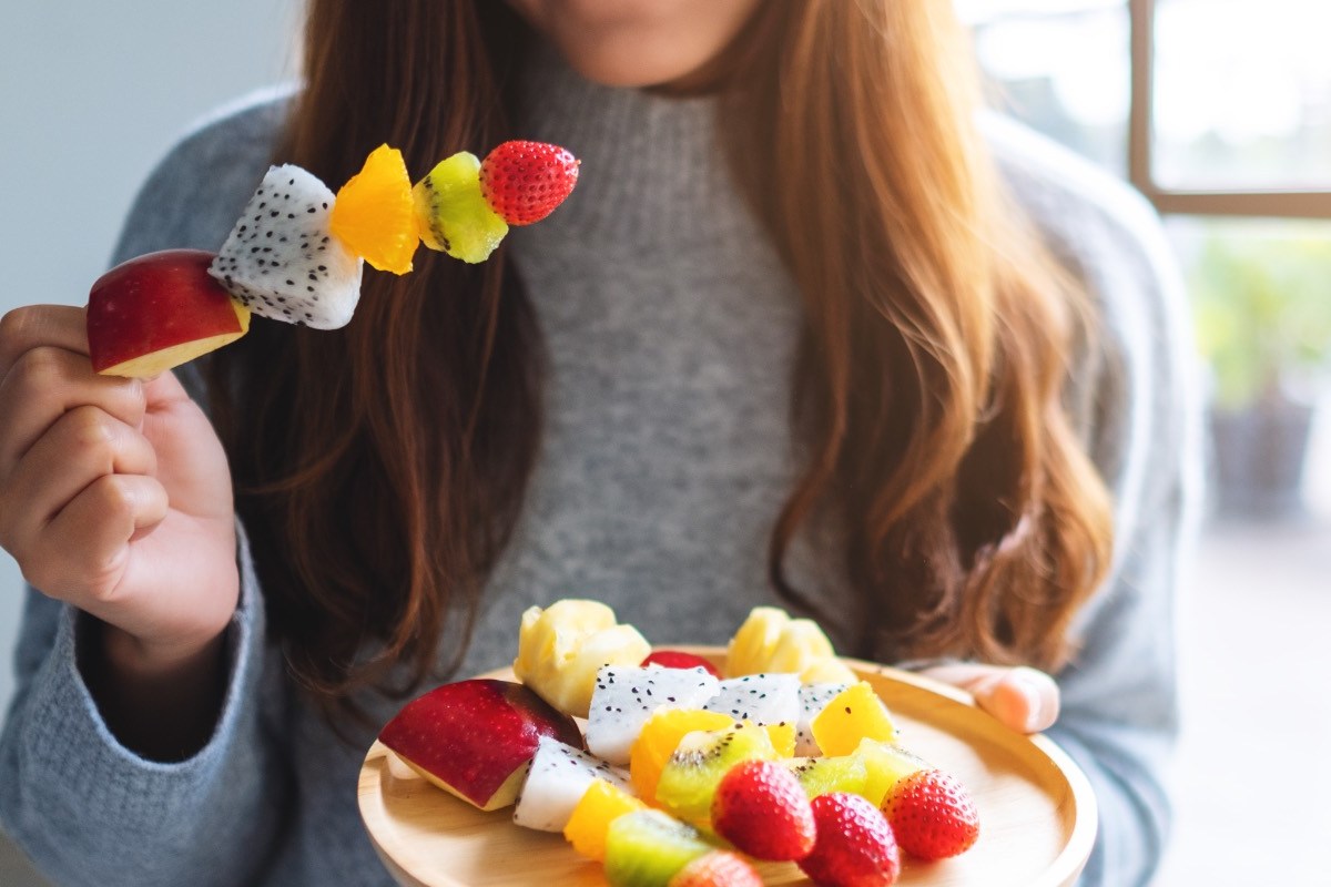 Obst am Abend essen: Eine Frau hält einen Obstspieß in der rechten und einen Teller mit Obstspießen in de rlinken Hand.