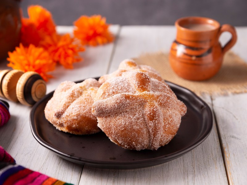 Pan de Muerto auf einem schwarzen Teller. Dahinter steht eine Tasse mit mexikanischer heißer Schokolade.