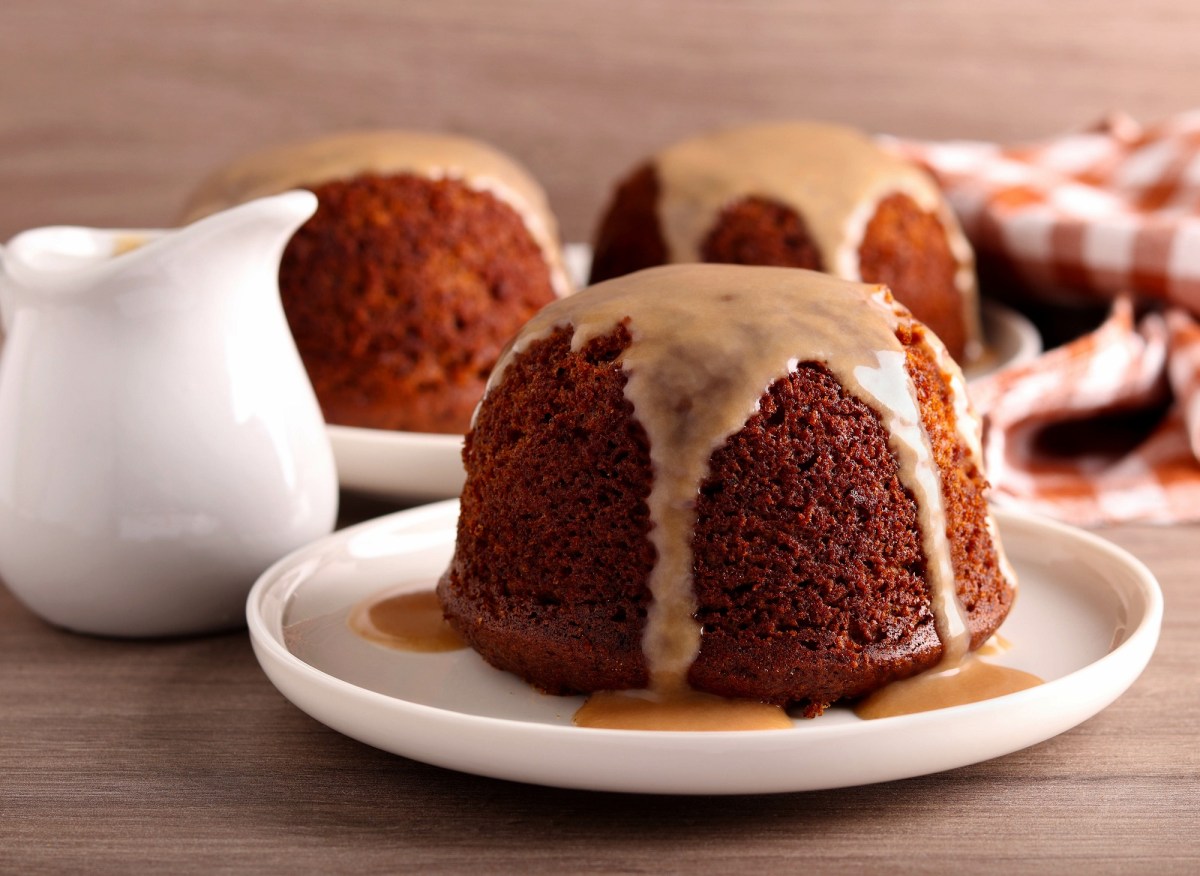 Sticky Toffee Pudding auf einem Teller, übergossen mit Karamellsoße. Daneben steht ein Kännchen mit Soße.