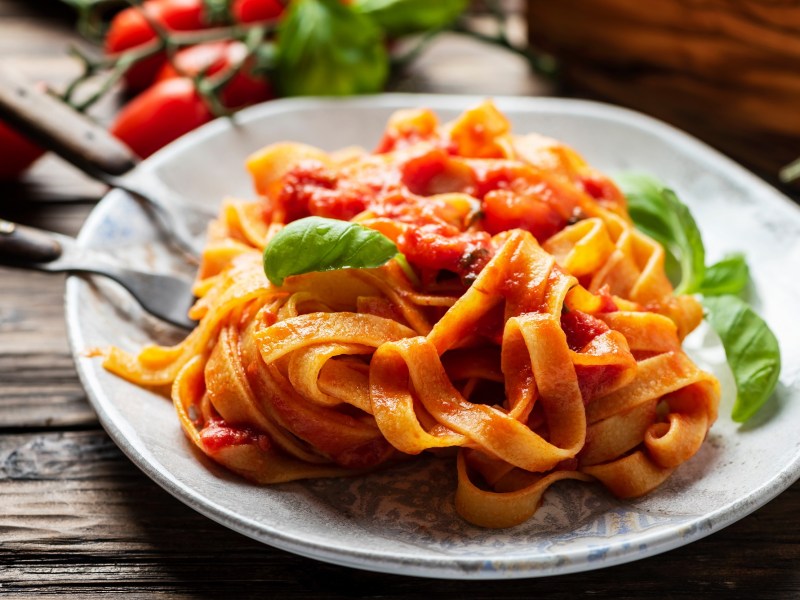 Ein Teller Tomaten-Creme-Pasta garniert mit Basilikum, daneben Besteck, Nahaufnahme.