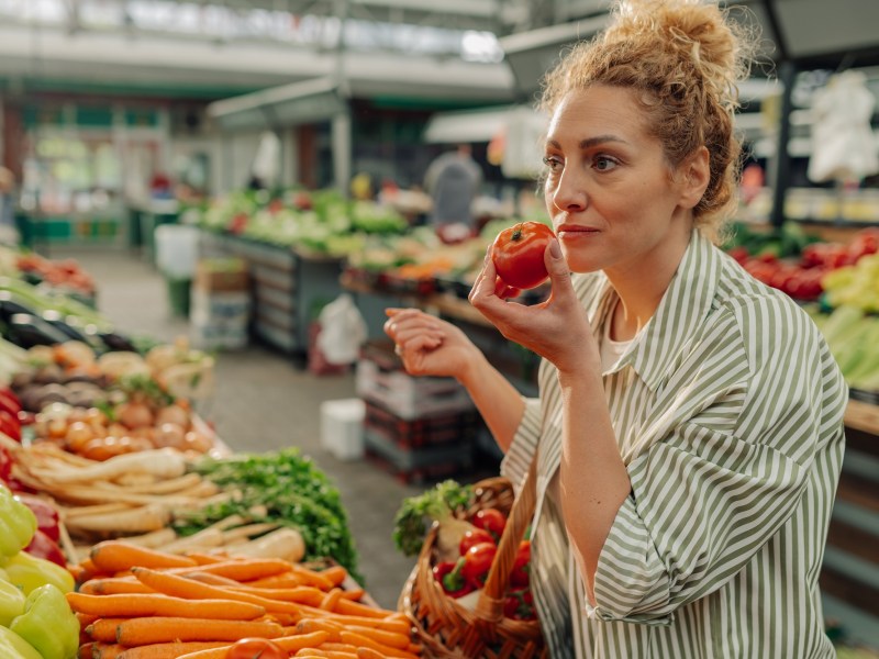 Vegane Mythen: eine Frau steht am GemÃ¼sestand und hÃ¤lt eine Tomate in der einen und einen Einkaufskorb in der anderen Hand.